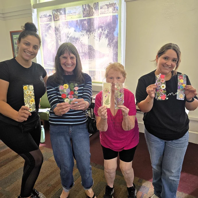 Four women standing indoors smiling, holding colorful bookmarks decorated with pressed flowers.