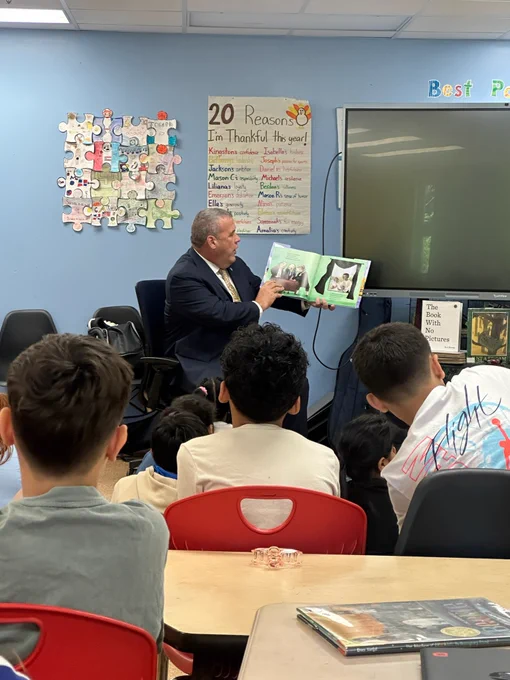 Secretary Amore reads a children's book aloud to a group of attentive elementary students seated in a classroom.