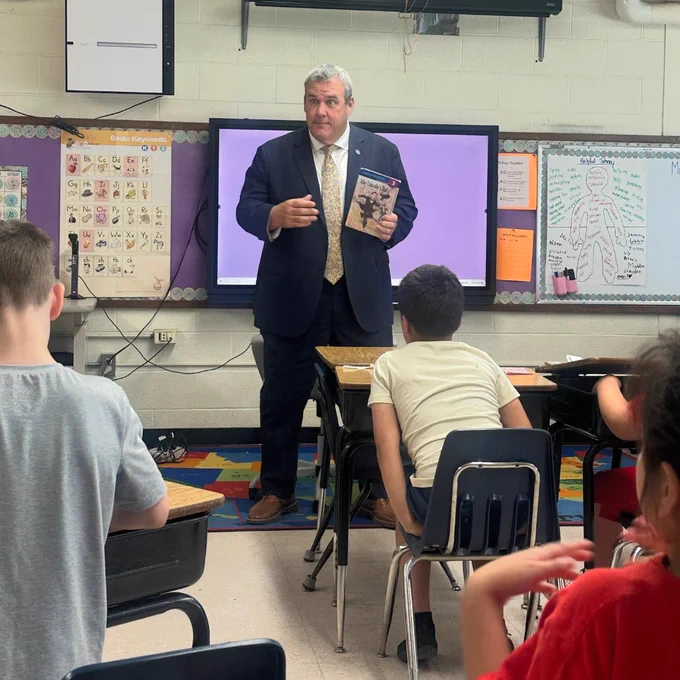 Secretary Amore holding a book in front of a group of attentive elementary students seated in a classroom.