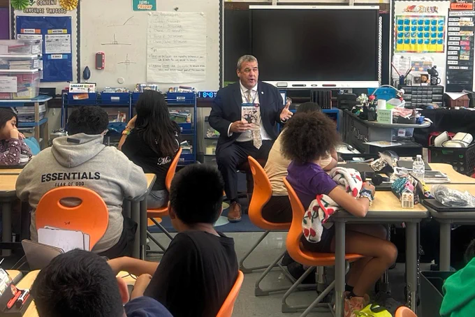 Secretary Amore holding a book in front of a group of attentive elementary students seated in a classroom.