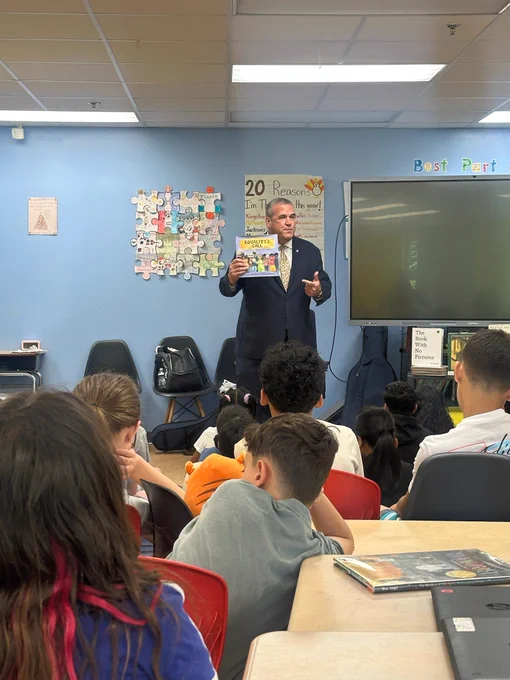 Secretary Amore holding a book in front of a group of attentive elementary students seated in a classroom.