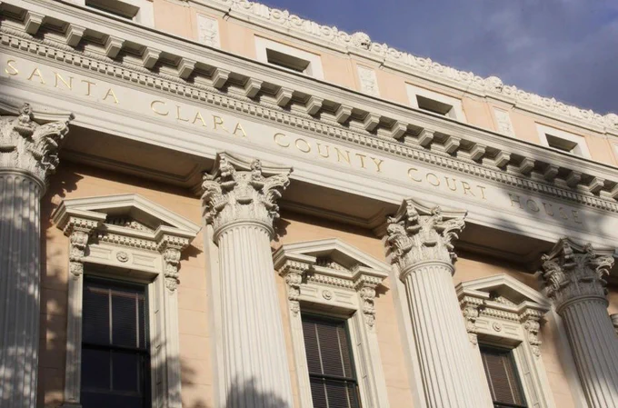 Exterior view of the historic Santa Clara County Courthouse, featuring tall Corinthian columns and ornate architectural details. The words “Santa Clara County Court House” are engraved across the upper facade beneath a clear blue sky.