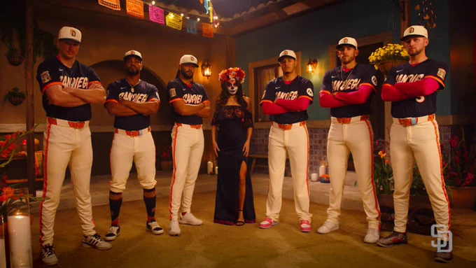A group of baseball players from the San Diego Padres stands in a room decorated for Día de los Muertos. They are dressed in uniforms. A person in a traditional Día de los Muertos outfit and face paint is at the center. The setting includes vibrant papel picado, candles, and flowers.