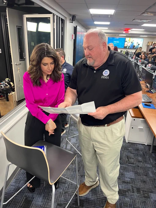 Senior Official Performing the Duties of Administrator Bob Fenton in a black polo discusses documents with Nicole Sganga in a bright pink blouse inside a busy office environment.