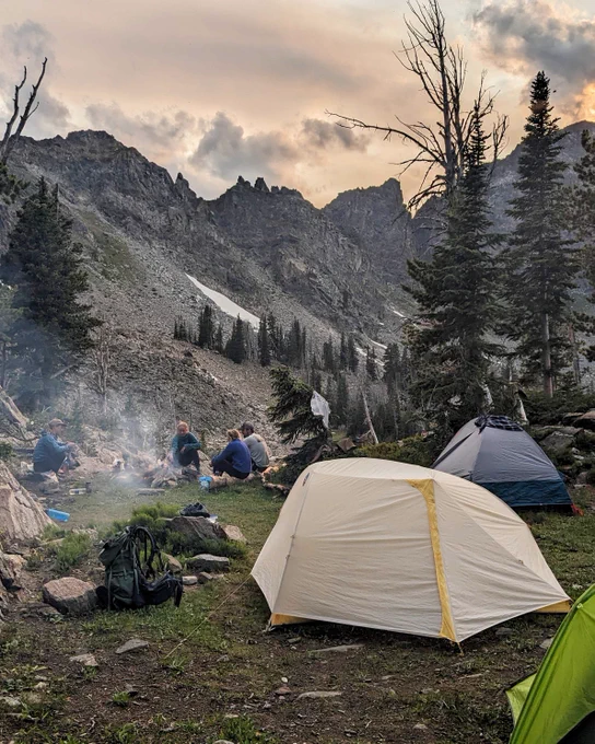 four people sitting around campfire with mountains in the background and tents in the foreground. 