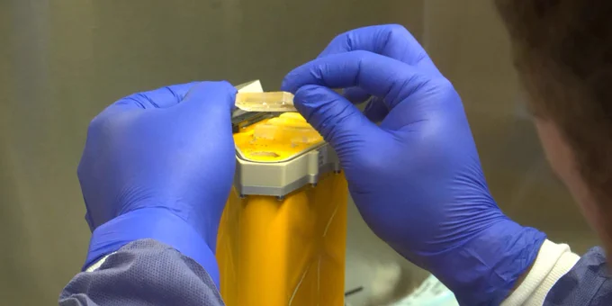 A close-up view of a person wearing blue gloves carefully handling a NASA's AVATAR investigation above a yellow scientific instrument in a laboratory setting.
