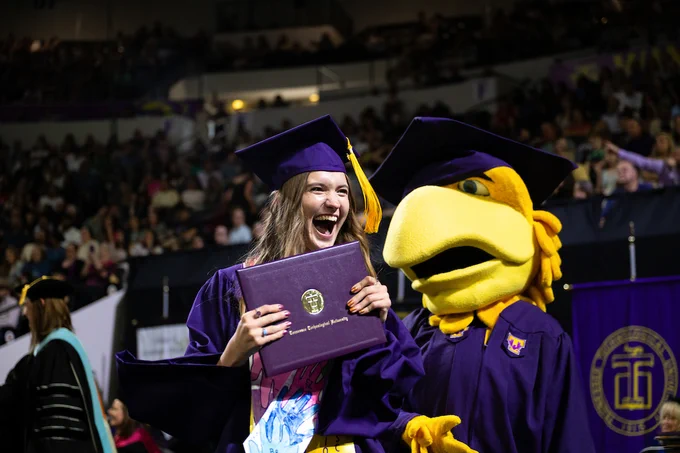 A graduate in a purple cap and gown smiles widely while holding a diploma cover. Next to the graduate is a large yellow eagle mascot, also in graduation attire. A crowd and university banners are visible in the background.