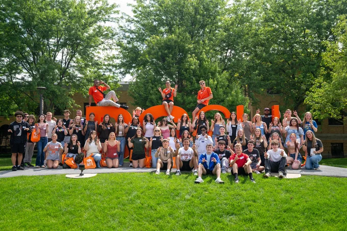 A group of people poses in front of large orange letters spelling "BGSU" on a sunny day. The background features green trees, and some individuals are sitting on the letters.