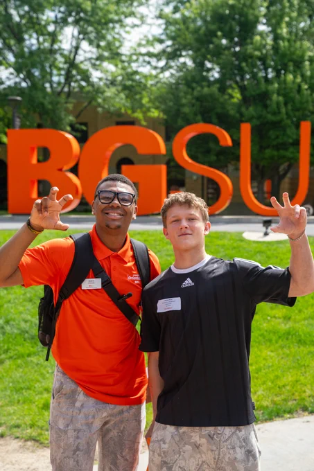 Two people standing in front of large orange letters spelling "BGSU" outside. Both are smiling and making a gesture with their hands. They are wearing casual clothes and name tags. Green grass and trees are in the background.