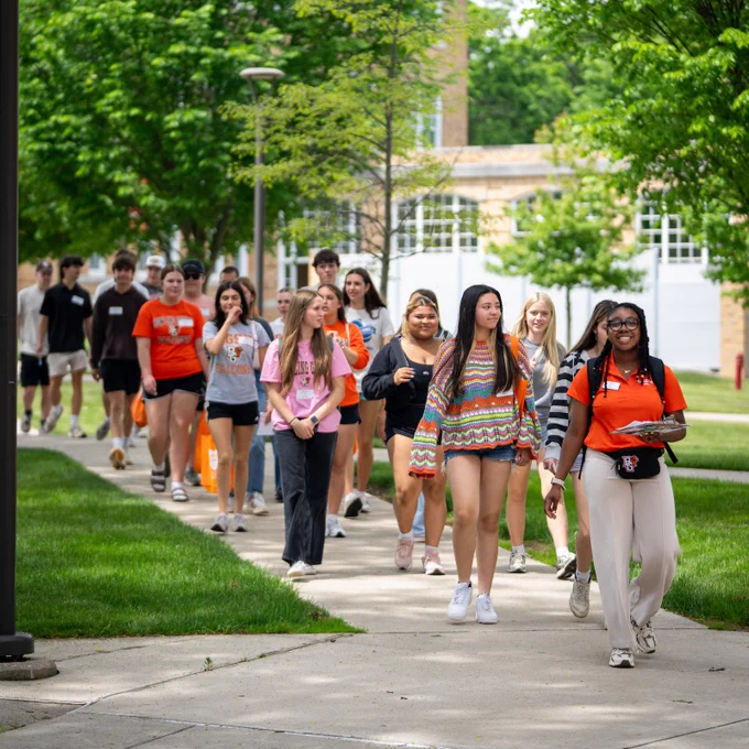 A diverse group of people walking together on a college campus sidewalk, surrounded by green grass and trees. They are wearing casual summer clothing and carrying bags and backpacks.