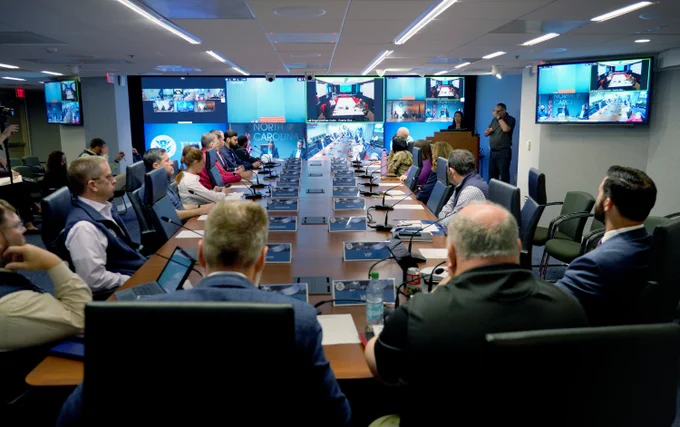 Interagency officials seated around a conference table in the National Response Coordination Center at FEMA headquarters in Washington, DC with a virtual call displayed on a screen. 