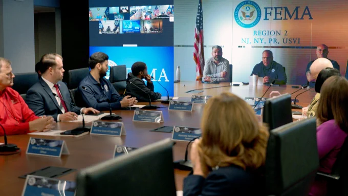 Interagency officials seated around a conference table in the National Response Coordination Center at FEMA headquarters in Washington, DC with a virtual call displayed on a screen.