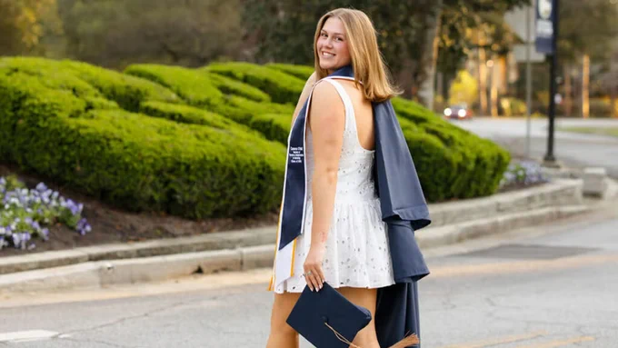 A person in a white dress and blue graduation gown looks back while walking on a road lined with greenery. The GSU bushes of Georgia Southern University are visible in the background. They hold a graduation cap and seem to be in a celebratory mood outdoors.