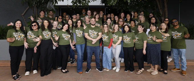 About 40 employees take a group photo in matching t-shirts that are olive green and say More Good Days Together. 