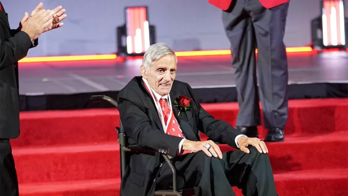 Bob Guzzo in a wheelchair wearing a black suit and red tie, seated on a red carpeted stage with people applauding nearby.