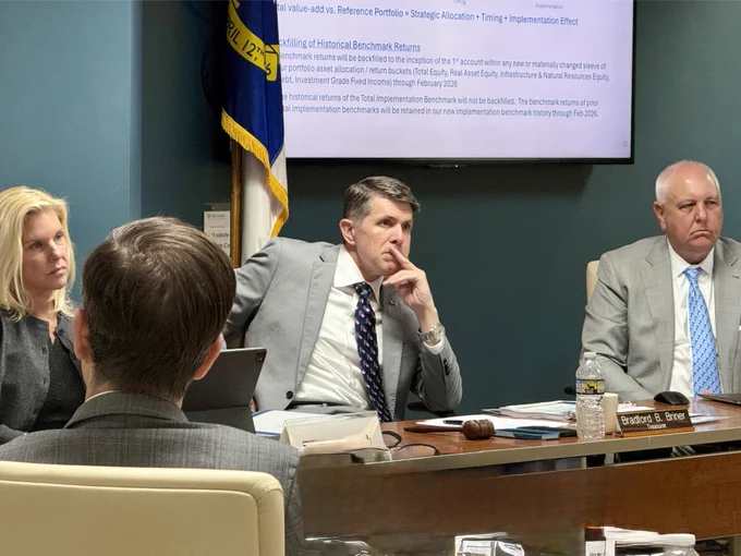 A group of people are seated around a conference table in a meeting room. There is a presentation slide projected on the wall with text discussing investment-related topics. An American flag and a state flag are visible in the background. Three people are attentively listening, and one person is taking notes. N.C. Treasurer's nameplate reading "Bradford B. Briner" is visible.