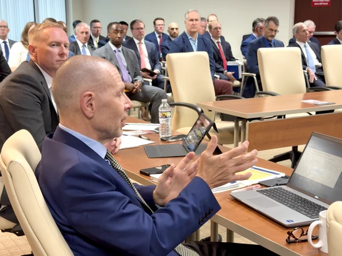 A group of people are seated in a conference room, attentively listening to a speaker. They are dressed in business attire, and each has a laptop in front of them. The setting appears formal, with mostly men in suits, engaged in the North Carolina Investment Authority meeting.