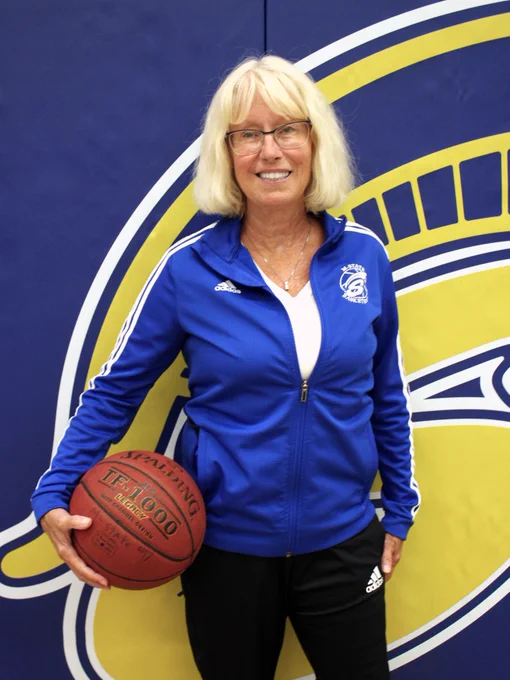 A person in a blue Adidas jacket holds a basketball, standing in front of a wall with a logo featuring a stylized Spartan helmet.
