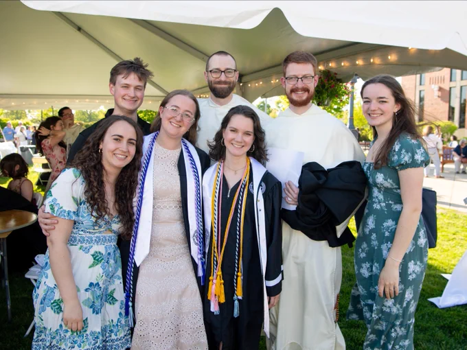 A group of seven people smiling under a white canopy at an outdoor event. Some are wearing graduation attire with cords and stoles. They are standing on a grassy area with a crowd and trees visible in the background.