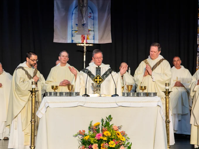 A group of clergy members in white robes stand behind an altar adorned with chalices and floral arrangements. A crucifix and a religious tapestry are visible in the background.
