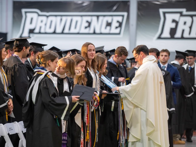A group of graduates in caps and gowns stands in line at a commencement ceremony. They are receiving diplomas from a figure in a white robe. "Providence" is prominently displayed on banners in the background.