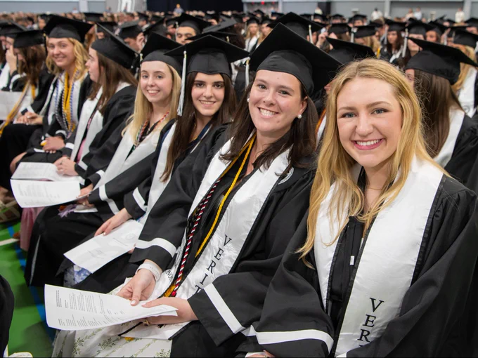 A group of graduates in caps and gowns sit together, smiling. They are holding programs, and their stoles say "VERITAS." The setting appears to be a graduation ceremony in a large hall with numerous other graduates in the background.