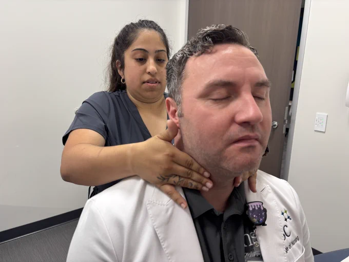 A person in a gray scrubs is examining the neck and shoulders of a person in a white coat. The person in the white coat has closed eyes and appears relaxed. They are standing in an exam room setting with white walls and a wooden door in the background.