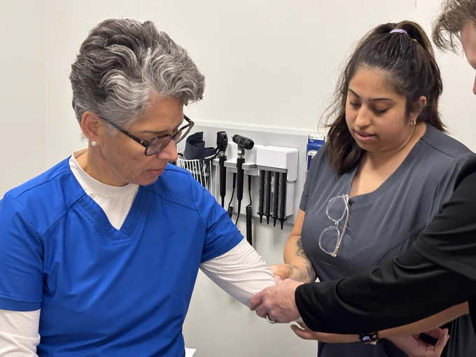 A healthcare worker in blue scrubs sits while a medical professional assists with checking or wrapping their forearm. Medical equipment hangs on the wall in the background.