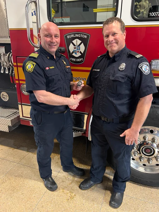 Deputy Chief and newly promoted Captain stand in fire department uniforms are shaking hands in front of a Burlington Fire Department truck.