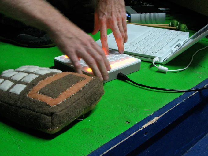 A big plushy soft calculator, Brian's hands on a 40h monome grid unit with USB cable sticking out, a white iBook G4, on the green floor of Etsy HQ in Brooklyn, August 2007.