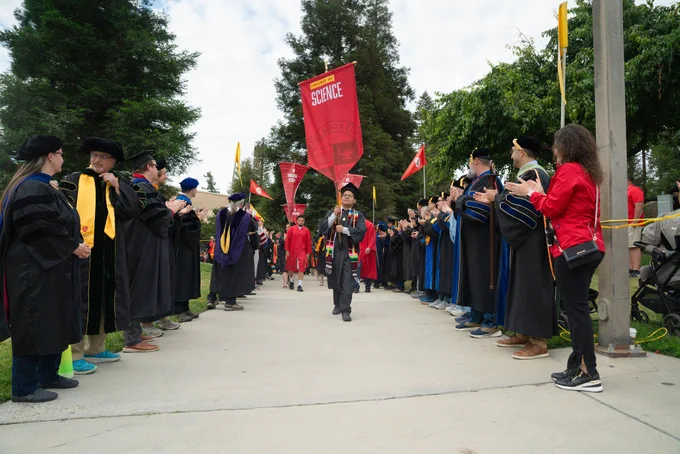 A graduate in a cap and gown walks through a faculty honor guard during a commencement ceremony, under a red "College of Science" banner. The atmosphere is celebratory.