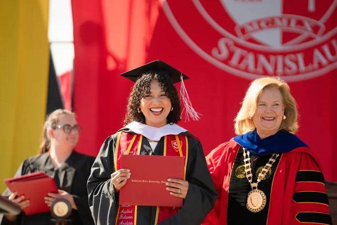 A graduate in a cap and gown joyfully holds her diploma, flanked by a smiling faculty member in regalia. A large red banner is visible in the background.