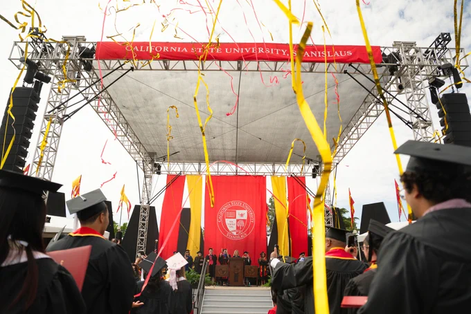 Graduates in caps and gowns face a stage with a red banner at California State University Stanislaus, with colorful streamers in the air. Festive atmosphere.