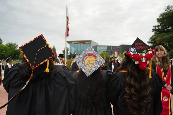 Three graduates in caps and gowns face away, showcasing decorated caps with a Trojan helmet, red flowers, and tassels, embodying jubilant celebration.