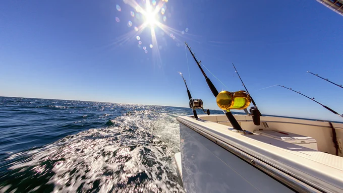 A boat travels across a sunlit ocean with several fishing rods mounted on its side. The sky is clear, and the sun shines brightly, reflecting off the waves.