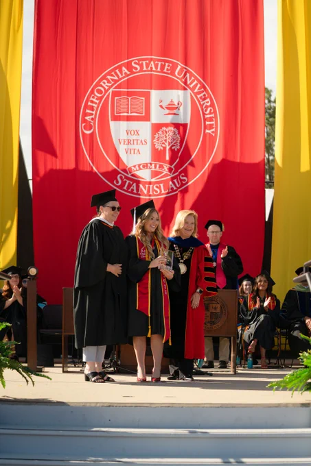 A graduate in cap and gown stands on stage at a graduation ceremony, holding a diploma. Faculty in academic regalia stand nearby, with a large red and gold California State University Stanislaus banner in the background. The scene conveys achievement and celebration.