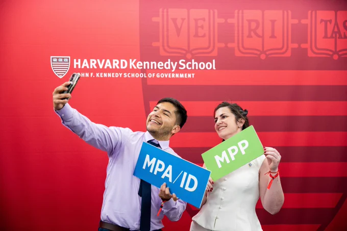 Twos students, one holding a blue MPA/ID sign and one holding a green MPP sign, taking a selfie.