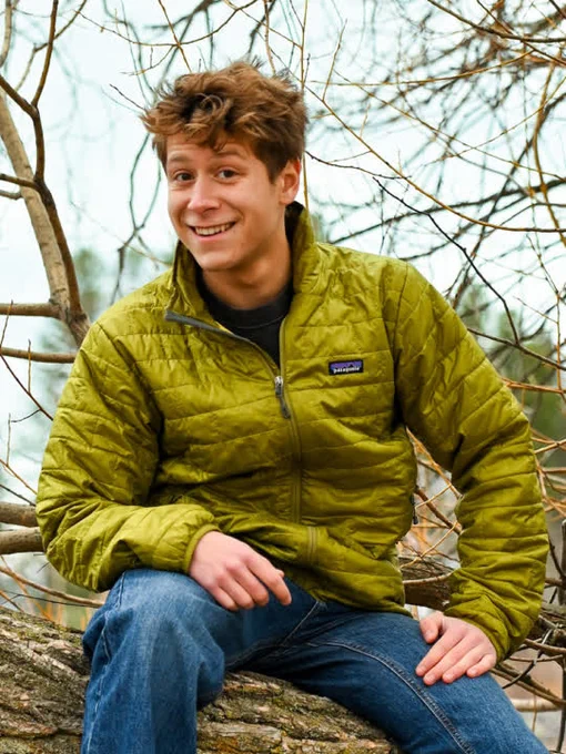 Noah Hamilton-Dixon sits on a tree branch, smiling and wearing a green coat