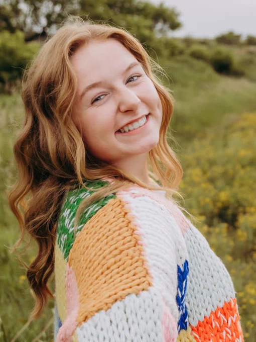 Rhyan Jenson wears a colorful sweater and smiles for the camera in front of a background of green grass and trees