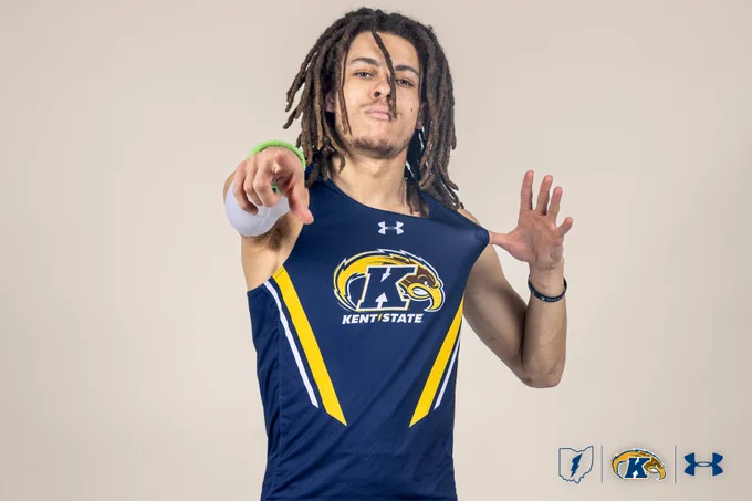 A person in a Kent State athletic jersey poses confidently, pointing towards the camera. The jersey displays the Kent State Golden Flashes logo, and the Under Armour logo is visible. The background is plain, and additional logos are at the bottom, including a feather, the Kent State emblem, and the Under Armour symbol.
