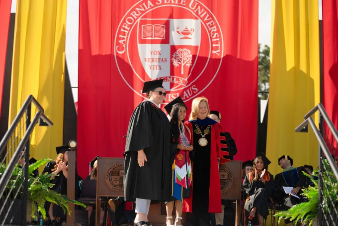 Graduates in caps and gowns stand on stage at a university ceremony. A large red banner with a white seal is in the background. Audience members sit nearby.