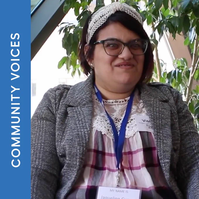 Smiling woman wearing glasses, lace headband, blazer and blue lanyard with badge, seated by plants; Community Voices banner on the left.