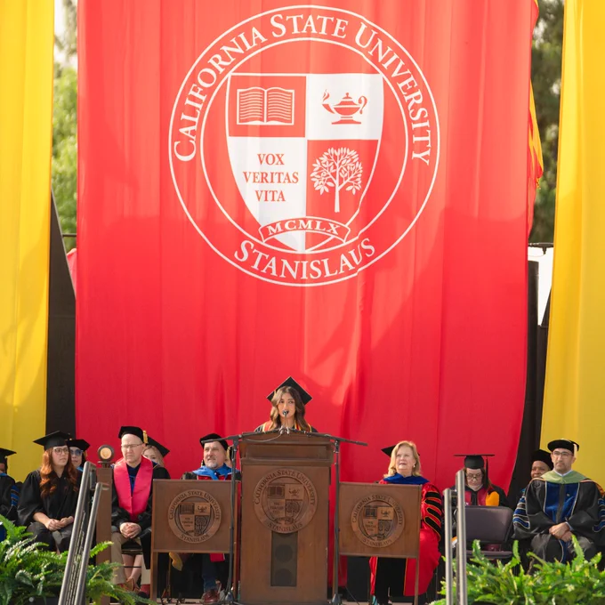 Graduation ceremony with a speaker at a podium wearing a cap and gown. Faculty in academic regalia sit behind her, with a vibrant red backdrop.