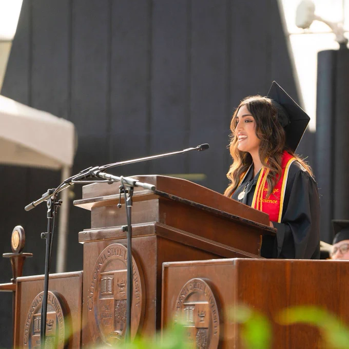 A smiling graduate in a cap and gown speaks at a podium during a sunny outdoor ceremony. She's wearing a red and gold stole, exuding pride and joy.