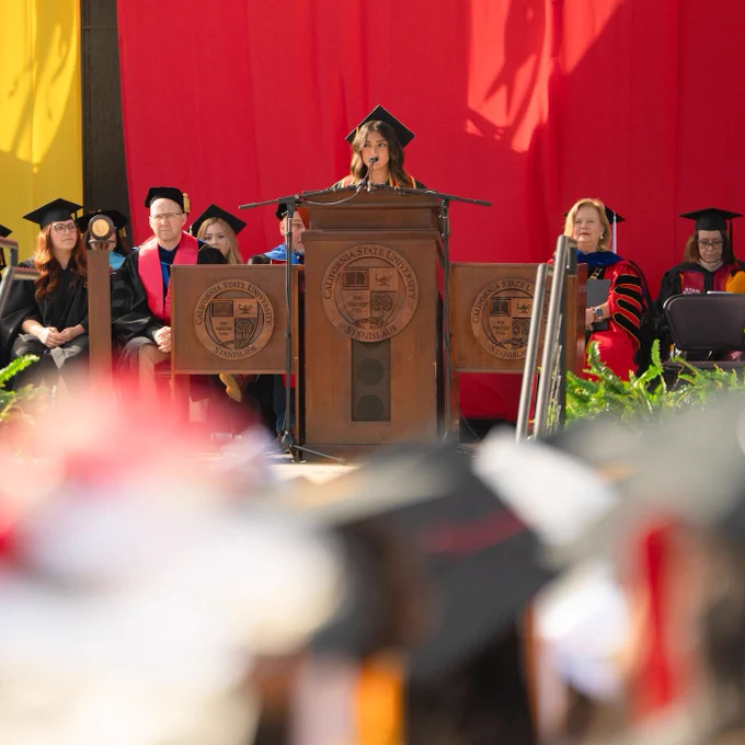 Graduation ceremony with a speaker at a podium wearing a cap and gown. Faculty in academic regalia sit behind her, with a vibrant red backdrop.