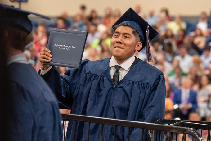 A young man in a blue graduation gown and mortarboard holds a diploma at a graduation ceremony surrounded by a crowd of people.