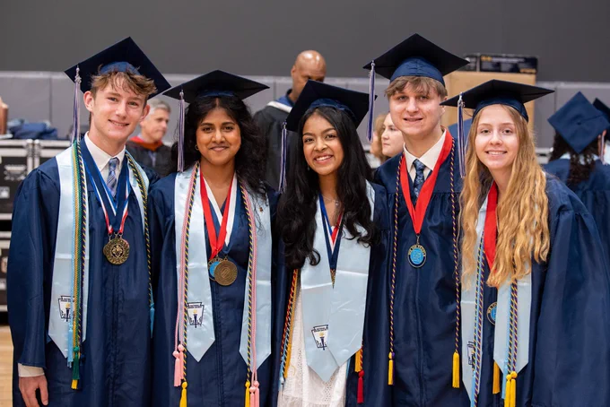A group of graduates in graduation gowns and mortarboards, some smiling and holding medals. The background appears to be an auditorium or stage.