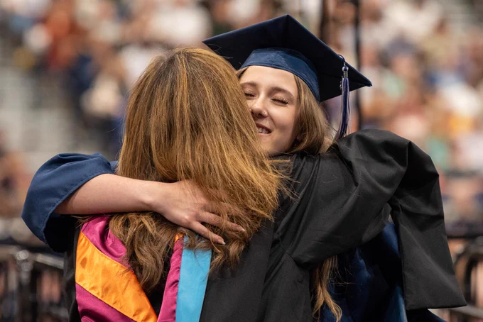 A graduate in a black gown and cap embraced by a person wearing colorful clothes, showing a celebratory moment.