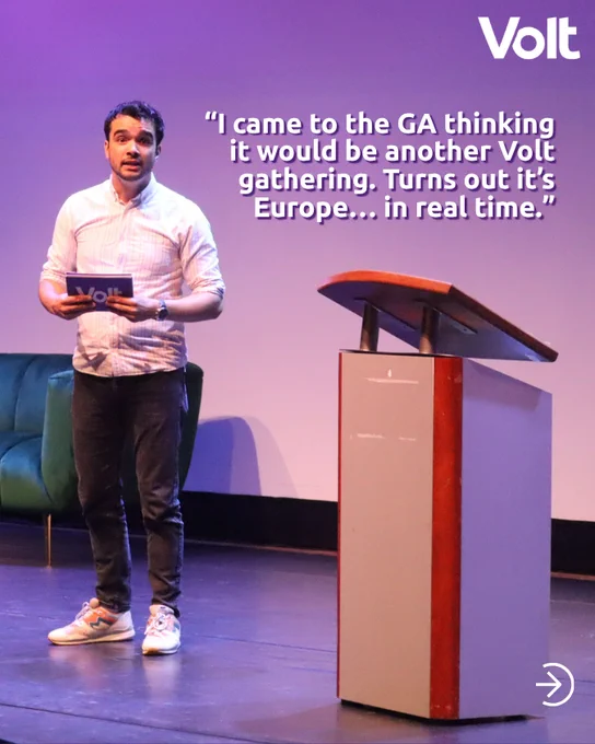 A man stands on a stage next to a wooden lectern, holding a Volt-branded booklet. He looks toward the audience with an engaged expression. Overlaid text reads: "I came to the GA thinking it would be another Volt gathering. Turns out it’s Europe... in real time." The Volt logo is in the top right corner.