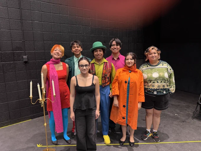 A group of seven individuals posing indoors against a black wall. They are dressed in colorful, eclectic clothing, with one standing near a candelabrum.
